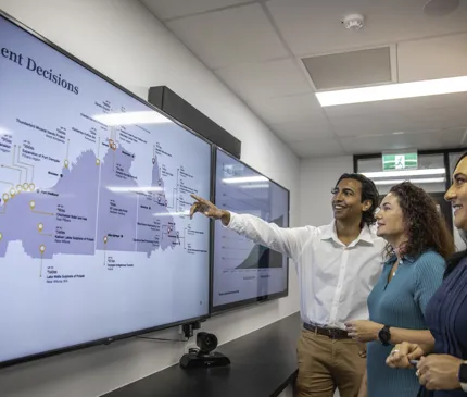 young man in white shirt and tan pants pointing at a screen with a map of our projects with a red haired woman wearing teal blue dress and another woman wearing dark blue dress